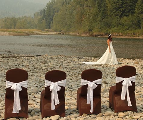 A row of four chairs covered in brown slipcovers tied with a large white bow are placed on a cobblestone river bank of the Elk River with a female dressed in a sleeveless wedding gown in the background.